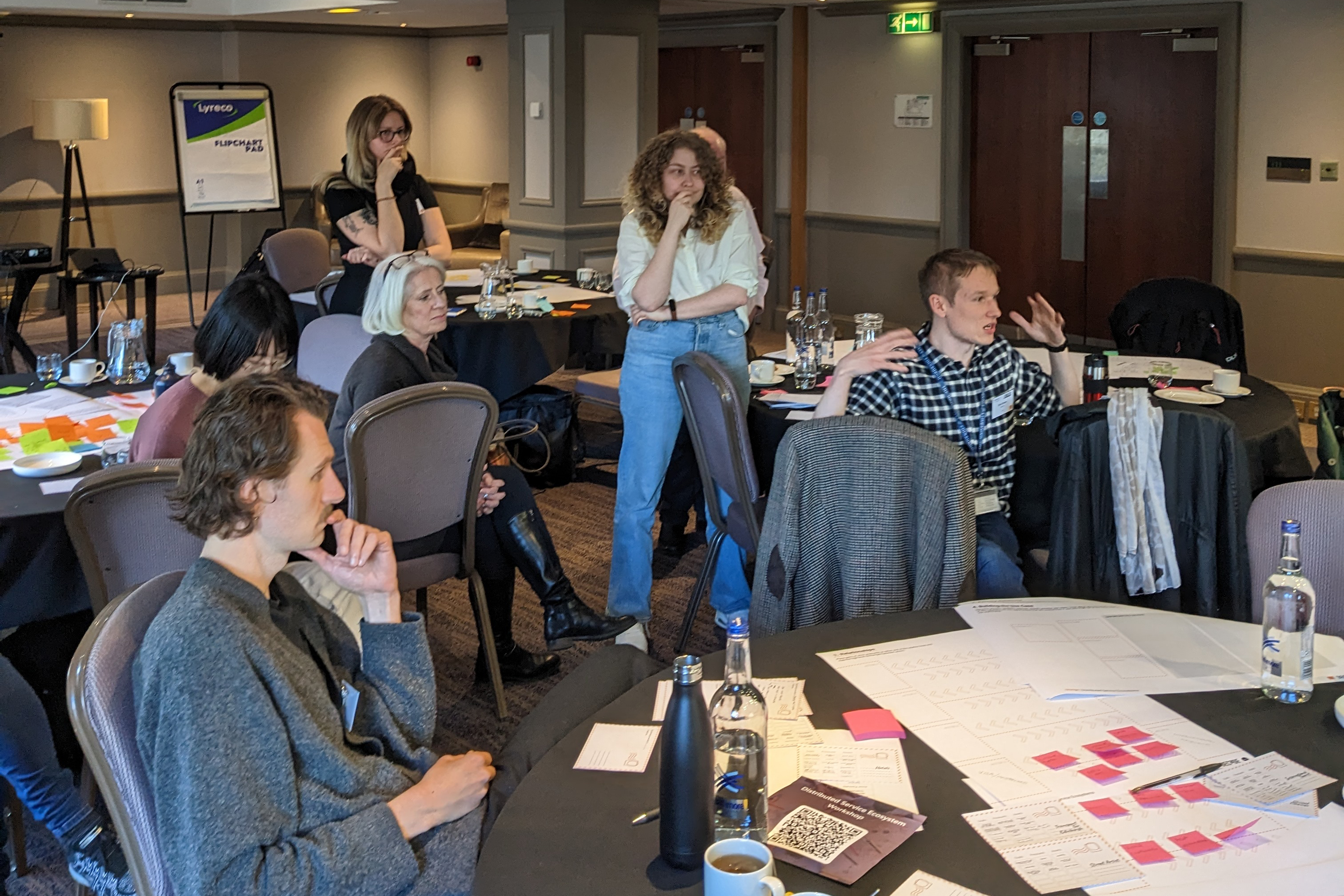 Participants collaborating in a workshop setting, engaging with printed worksheets and sticky notes on a table.